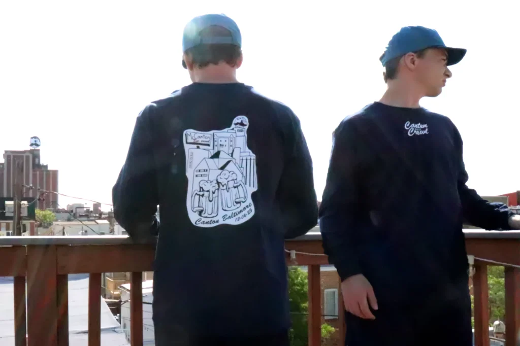 Rooftop morning photo of figures wearing the Front and back of a Bar Crawl t-shirt design. The shirt features the Natty Boh tower and the photograph features the actual natty boh tower in the background.